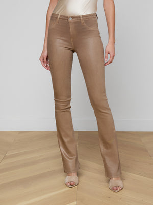 A woman stands indoors on light wooden flooring, wearing tan Selma Coated Bootcut Jeans, a light beige satin top, and open-toe heels. Her arms rest by her sides against a plain wall backdrop.