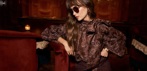 A woman wearing sunglasses and a brown paisley blouse sits on a red velvet chair in a dimly lit, elegant room with wood-paneled walls.