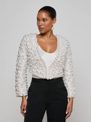 A woman models the L'AGENCE Belle Oversized Pearl Jacket, featuring hand-sewn faux pearls over a white top and high-waisted black pants. With sleek hair and gold earrings, she creates a striking look as she gazes off to the side.