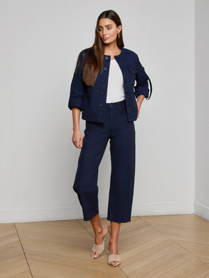 A woman with long brown hair and a neutral expression stands on a wooden floor indoors wearing the Anouk Jean Jacket in navy blue, styled with matching cropped pants, a white top, and beige heeled sandals.