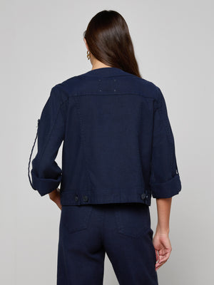 A person with long brown hair, seen from behind, wears the Anouk Jean Jacket— a collarless, relaxed-fit denim jacket with rolled-up sleeves, button details at the waistband, and matching dark blue pants.