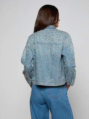 Seen from behind, a person with long brown hair wears the Vangie Embellished Jean Jacket featuring crystal details and a cropped high-low hem, paired with blue jeans against a plain gray background.