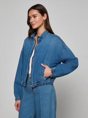 A woman wears the Dunia Jean Jacket over a white top and blue denim pants, standing with one hand in her pocket and angled slightly to the side against a plain light background.
