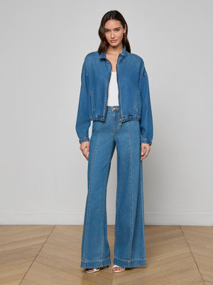 A woman with long, wavy hair stands indoors on a wooden floor, wearing the Dunia Jean Jacket, wide-leg high-waisted jeans, a white top, and sandals. She looks calm against a plain light-colored wall.