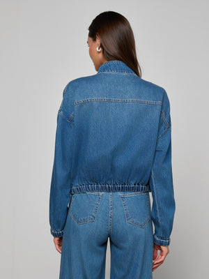 A woman with long brown hair, seen from behind, wears the Dunia Jean Jacket and blue denim jeans against a plain light gray background.