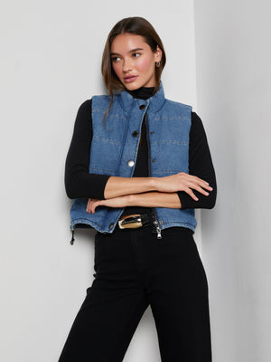 A woman with long brown hair wears the cropped, sleeveless indigo Rola Puffer Vest over a black long-sleeve top and pants, standing with arms crossed against a white wall and looking to the side.