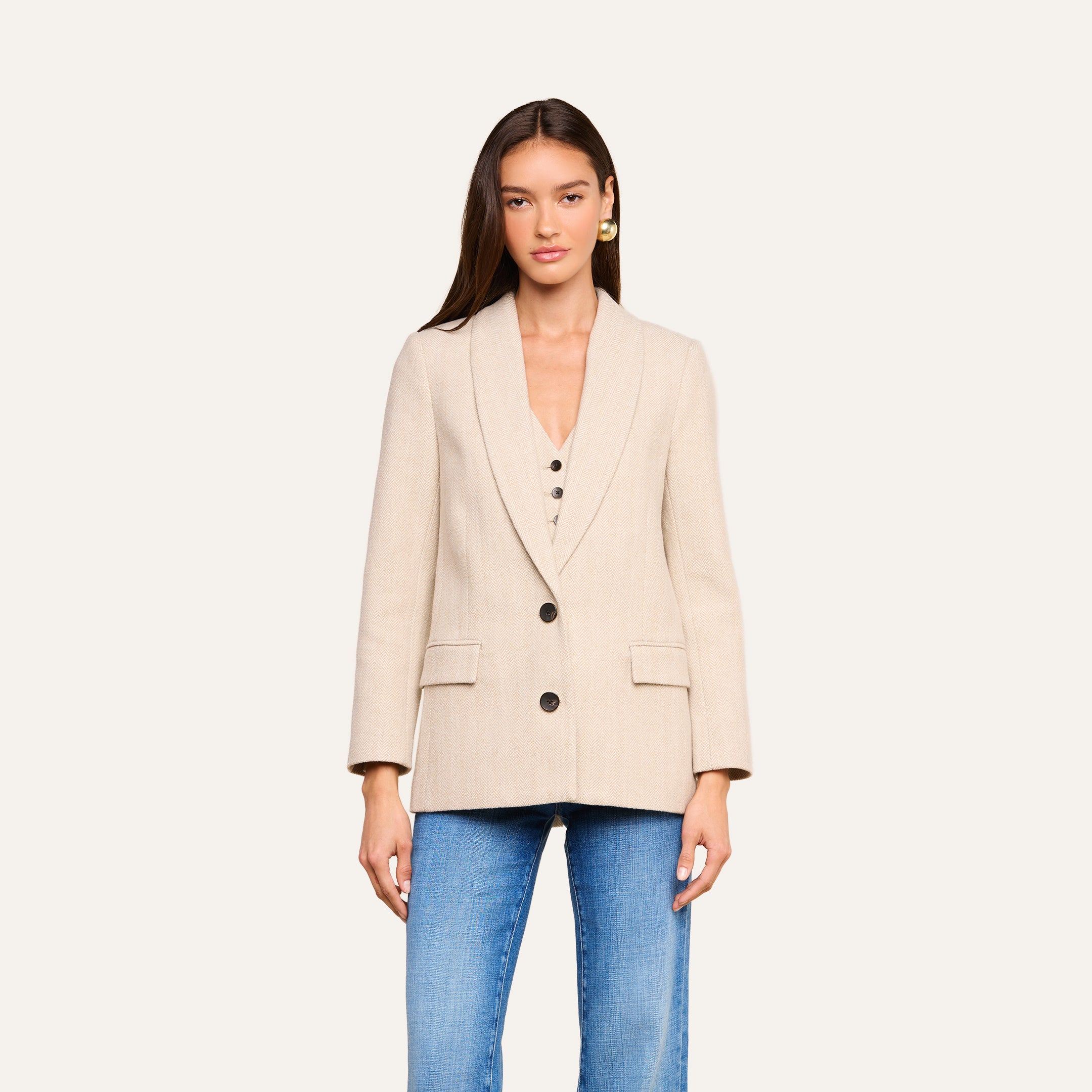 Una donna è in piedi, rivolta verso la fotocamera, su uno sfondo beige a tinta unita, incarnando l’eleganza senza tempo che spesso caratterizza la Settimana della Moda di New York, indossando l’Amelie Blazer — una giacca beige dal taglio aderente con bottoni neri — abbinata a jeans blu.