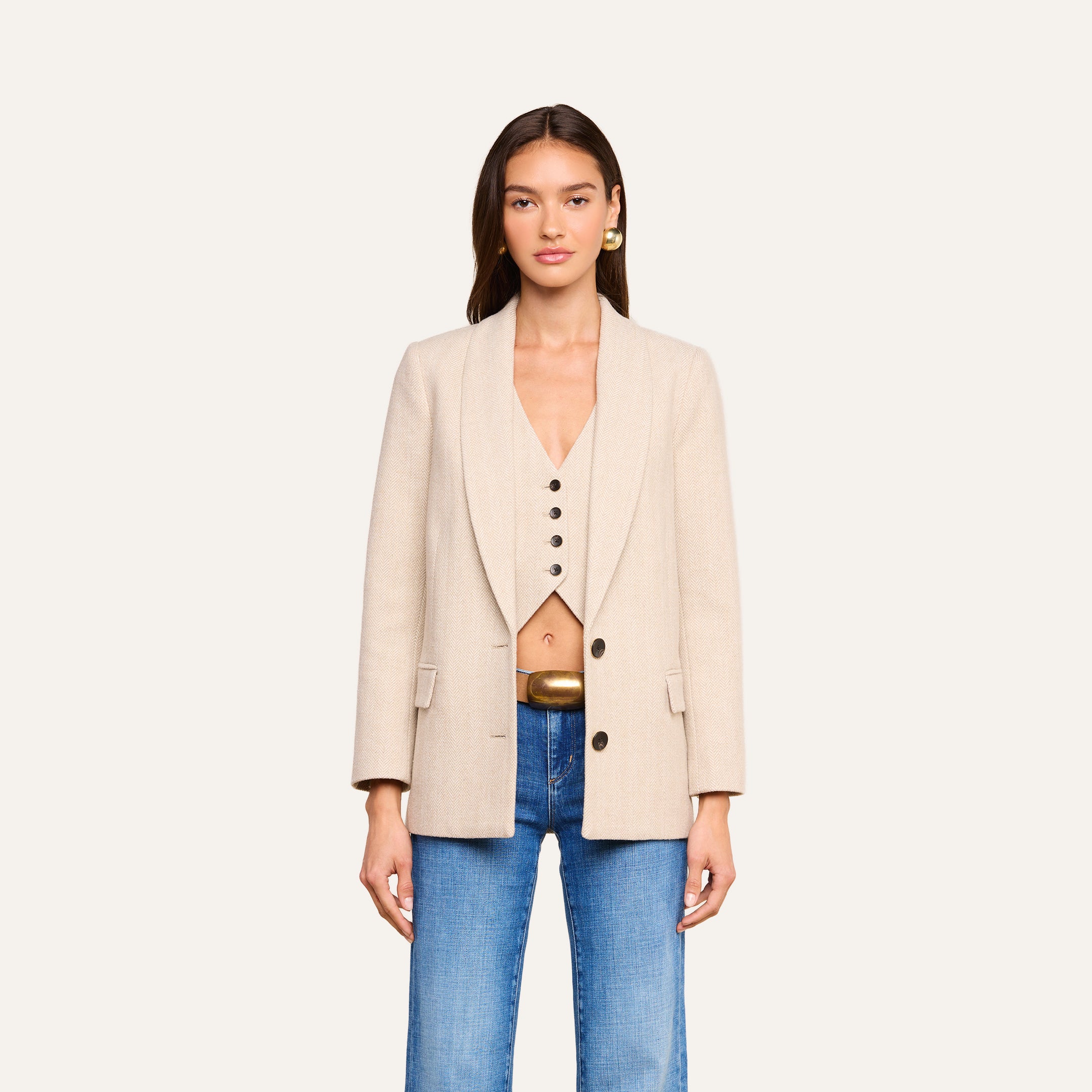 Una donna è in piedi, rivolta verso la fotocamera, e indossa il blazer Amelie beige dalla linea aderente sopra un gilet beige abbottonato che lascia scoperto l’ombelico, abbinato a jeans blu e orecchini dorati, il tutto su uno sfondo beige chiaro a tinta unita: un look che ricorda gli outfit chic visti alla Settimana della Moda di New York.