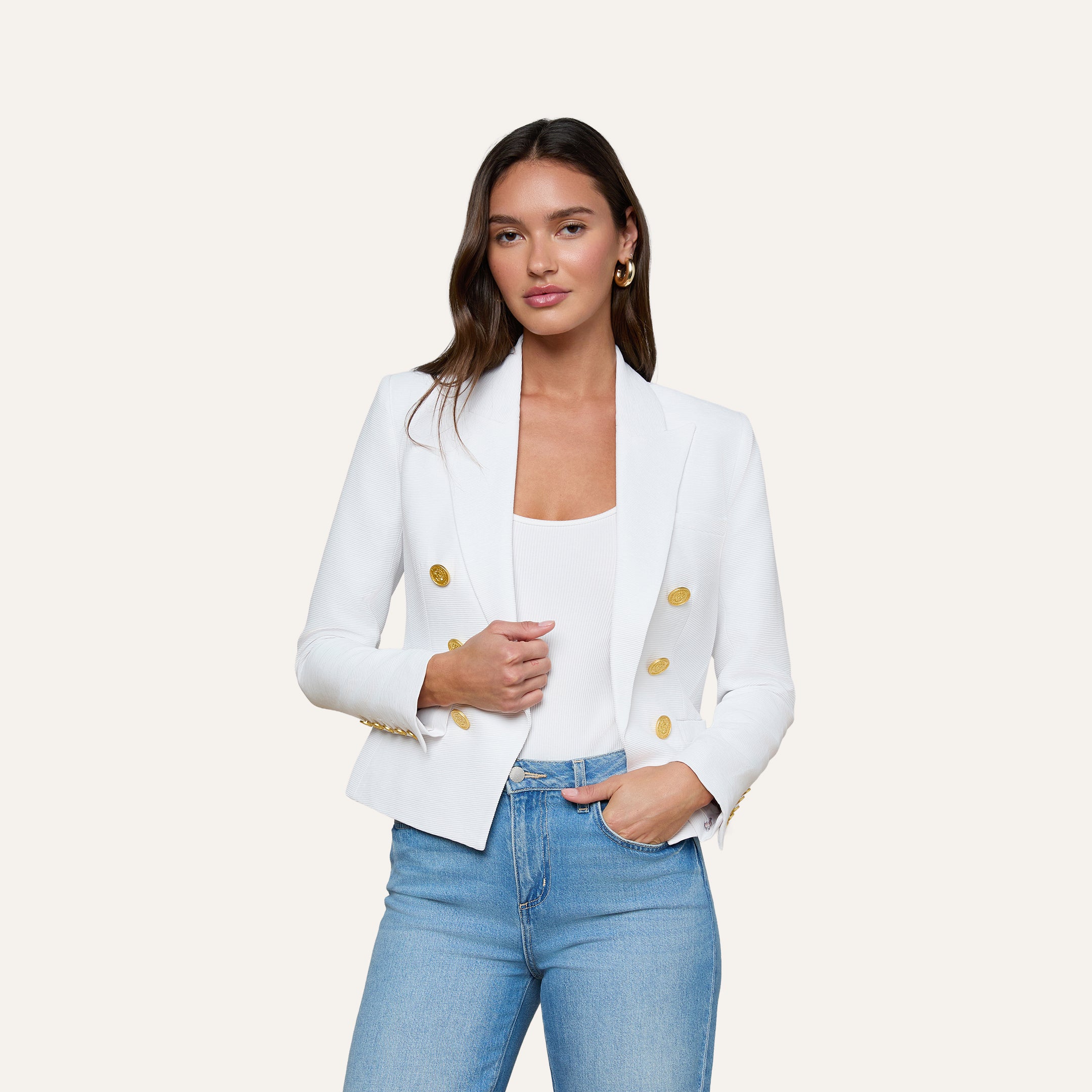 A woman stands against a plain light background with one hand in her pocket, wearing the Brooke Blazer—a white double-breasted blazer with gold buttons—paired with a white top and light blue jeans.