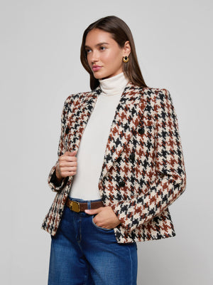 A woman stands against a plain background, wearing the Marie Houndstooth Tweed Blazer over a white turtleneck and blue jeans, accessorized with gold hoop earrings—one hand in her pocket and the other holding her blazer.