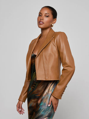 A woman models the Dulcie Leather Blazer, a tan jacket with whipstitch accents, layered over a vibrant peacock-print dress against a simple light gray backdrop, gazing confidently at the camera.