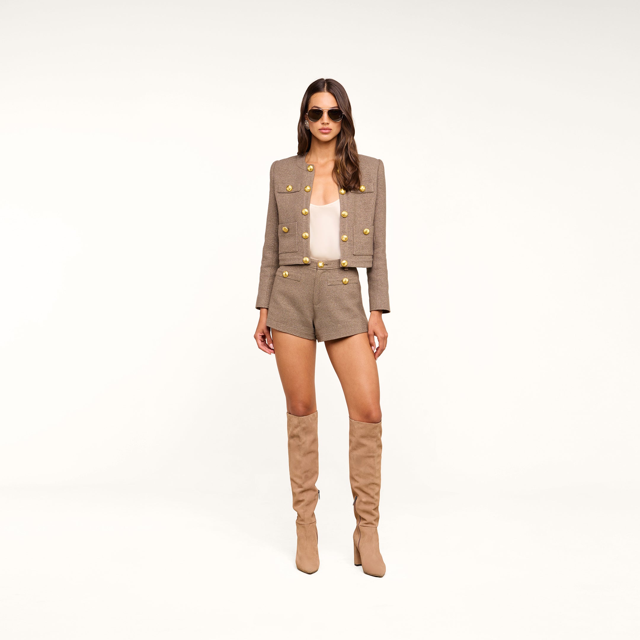 A woman stands against a plain white background wearing sunglasses, a tan jacket with gold buttons, the Dionne Tweed Short in a high-rise style, a white top, and knee-high tan boots.