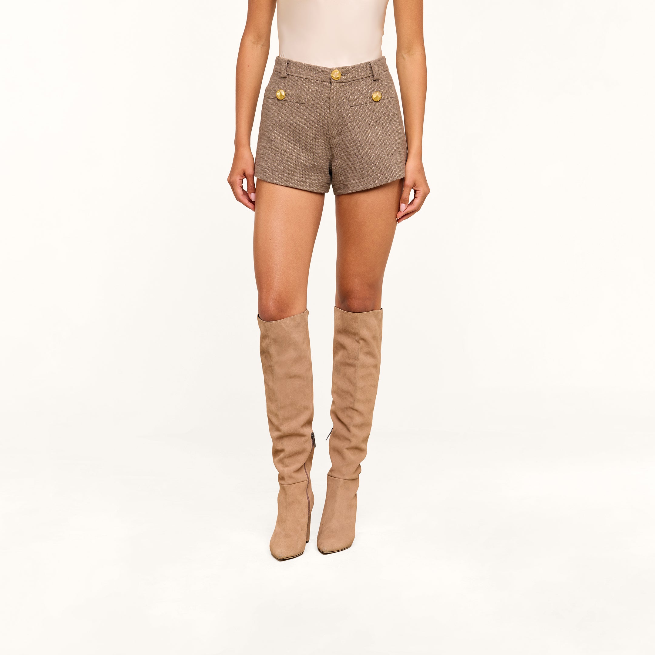 A person wearing a sleeveless beige top, the Dionne Tweed Short with gold buttons, and tall beige suede boots stands against a plain white background.