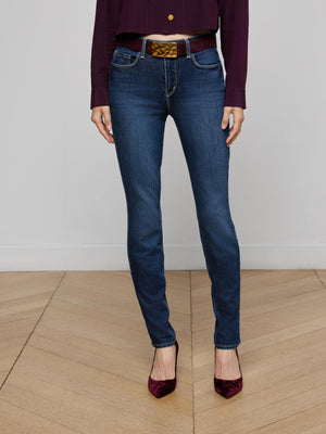Wearing L'AGENCE Marguerite Skinny Jeans in dark blue, a burgundy long-sleeve top, brown braided belt, and burgundy pointed-toe heels, a person stands on a wooden floor against a plain light-colored wall.
