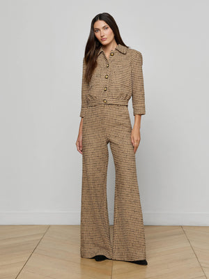 A woman with long dark hair and black shoes stands indoors against a plain wall and wooden floor, wearing Pilar Tweed Wide-Leg Pant in brown Italian windowpane tweed with a high rise, wide legs, large buttons, and a collar.