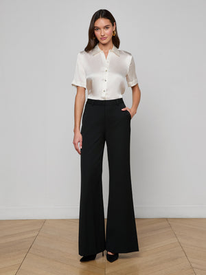 A woman stands indoors against a light wall, wearing a short-sleeve ivory satin blouse and the Pilar Wide-Leg Pant made from four-way stretch suiting. With her hands in her pockets, she faces forward in black heels.