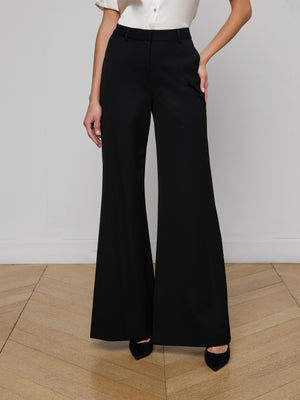 A person wears the Pilar Wide-Leg Pant in black with a white short-sleeve blouse, standing on a light wood floor with one hand on their hip. The high-waisted pants are crafted from four-way stretch suiting; their face is not visible.