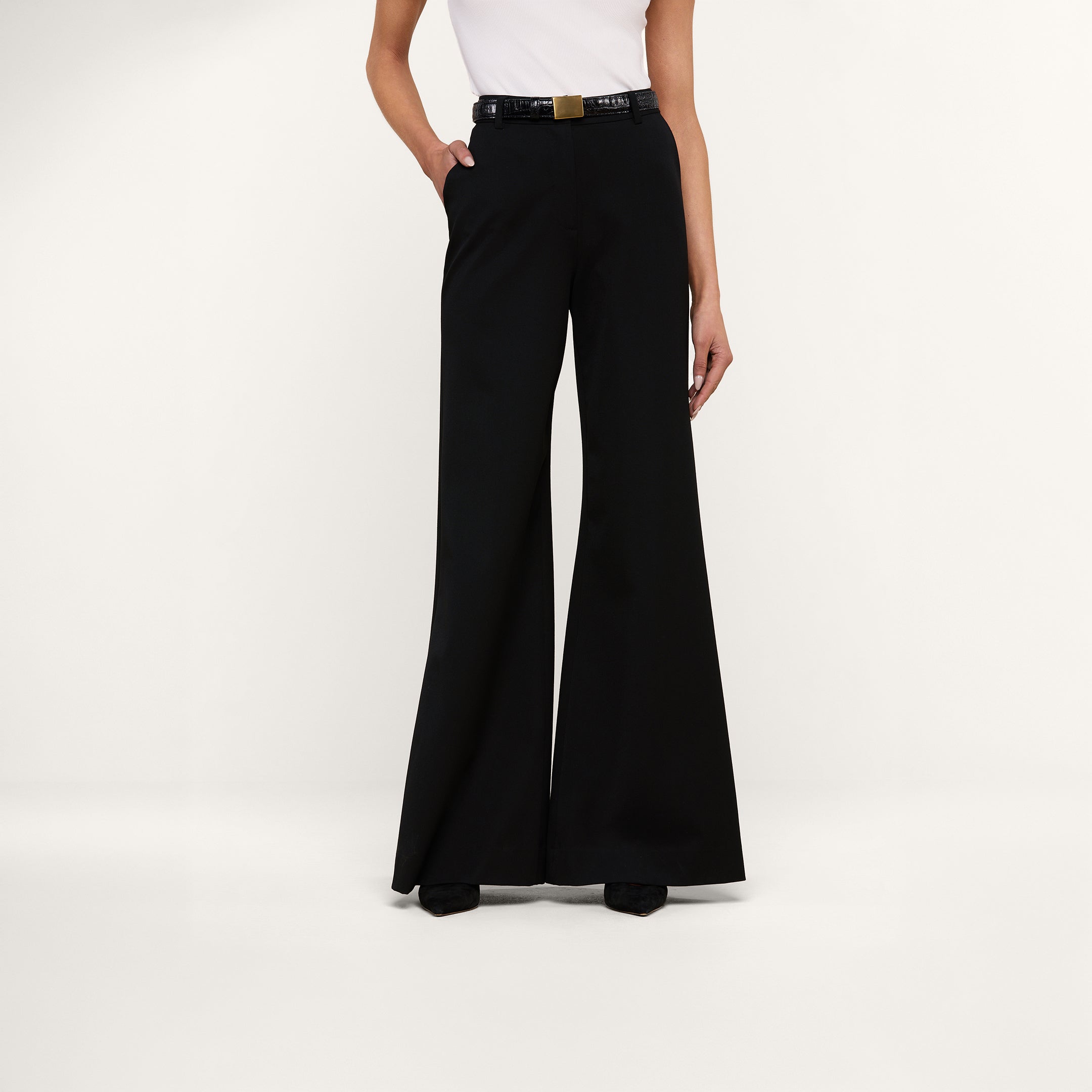 A person wearing the Pilar Wide-Leg Pant in black, crafted from four-way stretch suiting, paired with a white sleeveless top and a black belt with a gold buckle, stands against a plain white background.