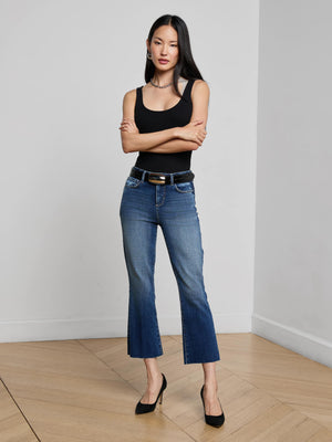 A woman with long dark hair, in a black tank top, belt, and heels, stands with arms crossed in a minimalist room. She finishes her look with L'AGENCE Kendra Cropped Flare Jeans in stretch denim.