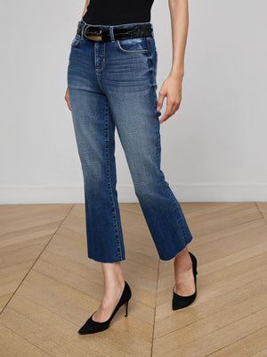 A person in L'AGENCE Kendra Cropped Flare Jean (dark wash) with a black top, black belt, and black high-heeled shoes stands on a light wood floor against a plain light-colored wall.