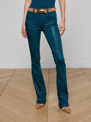 A person stands on a wooden floor wearing L'AGENCE Selma Coated Bootcut Jeans in teal with a brown belt and pointed brown shoes. The upper body is partially out of frame.