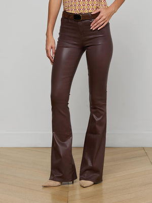 Wearing L'AGENCE Marty Coated Flare Jean in dark brown with a patterned top and beige pointed-toe heels, a person stands on a wooden floor against a white wall. The image is cropped at the shoulders.