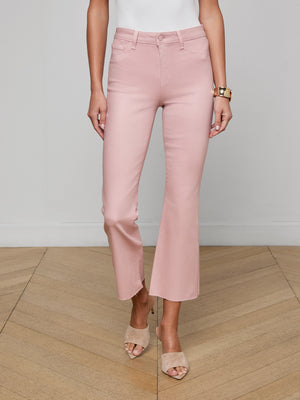 Wearing Kendra Coated Cropped Flare Jeans in light pink, a white sleeveless top, beige open-toe heeled sandals, and a gold bracelet, a person stands on light wood flooring against a plain light gray wall.