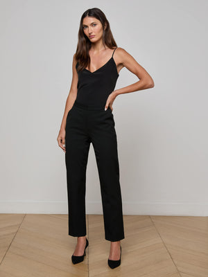 A woman with long brown hair wears a black sleeveless top and the Rebel Trouser, paired with black heels, standing on a light wooden floor against a plain white wall, one hand on her hip and the other relaxed by her side.