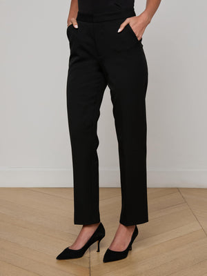 A woman stands with her hands in her pockets, wearing the Rebel Trouser—high-rise, straight-leg Italian stretch suiting—paired with black heels on a light wooden floor against a plain white wall.
