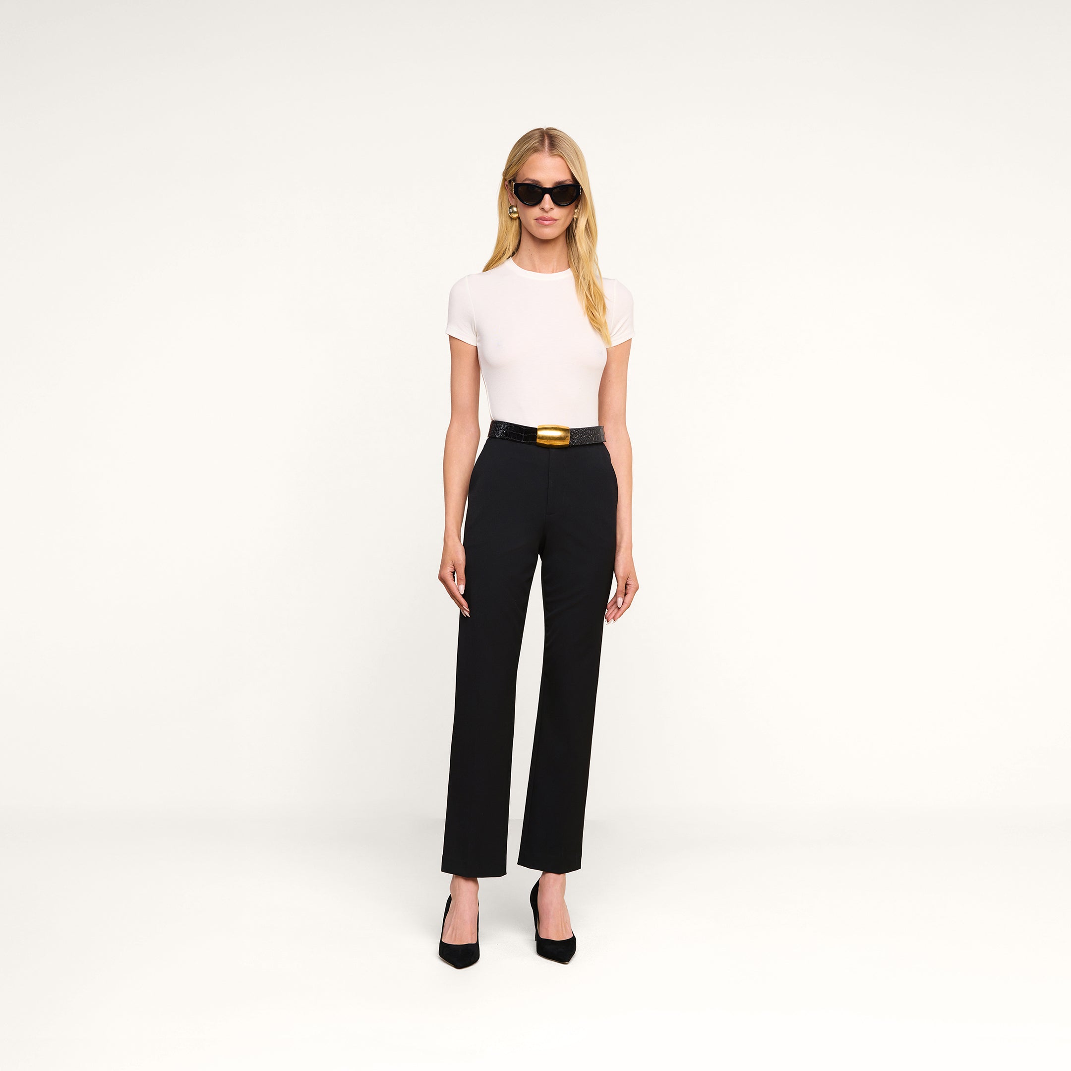 Une femme portant des lunettes de soleil, un haut blanc à manches courtes, le « Rebel Trouser » — un pantalon noir à taille haute et coupe droite, confectionné dans un tissu de costume italien extensible et agrémenté dune large ceinture — ainsi que des talons hauts noirs, se tient debout devant un fond beige.