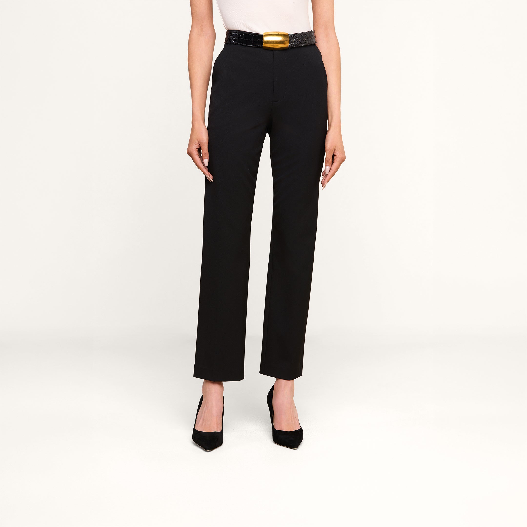 Une personne portant le pantalon « Rebel » — un pantalon noir à taille haute et coupe droite, confectionné dans un tissu de costume italien extensible —, associé à un haut blanc, une ceinture noire ornée dune grande boucle dorée et des escarpins noirs à bout pointu, se tient debout devant un fond beige.