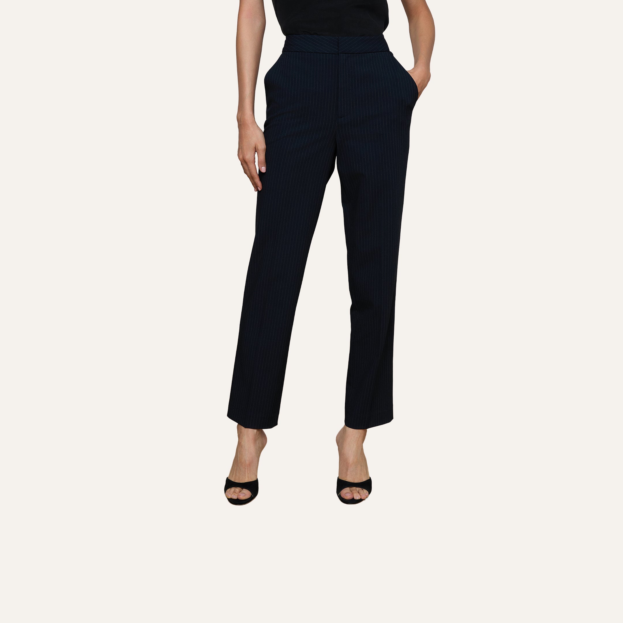 Wearing the Rebel Pinstriped Trouser in black high rise, paired with black open-toe heels and hands casually tucked into pockets, set against a plain light background. Upper body is partially out of frame.
