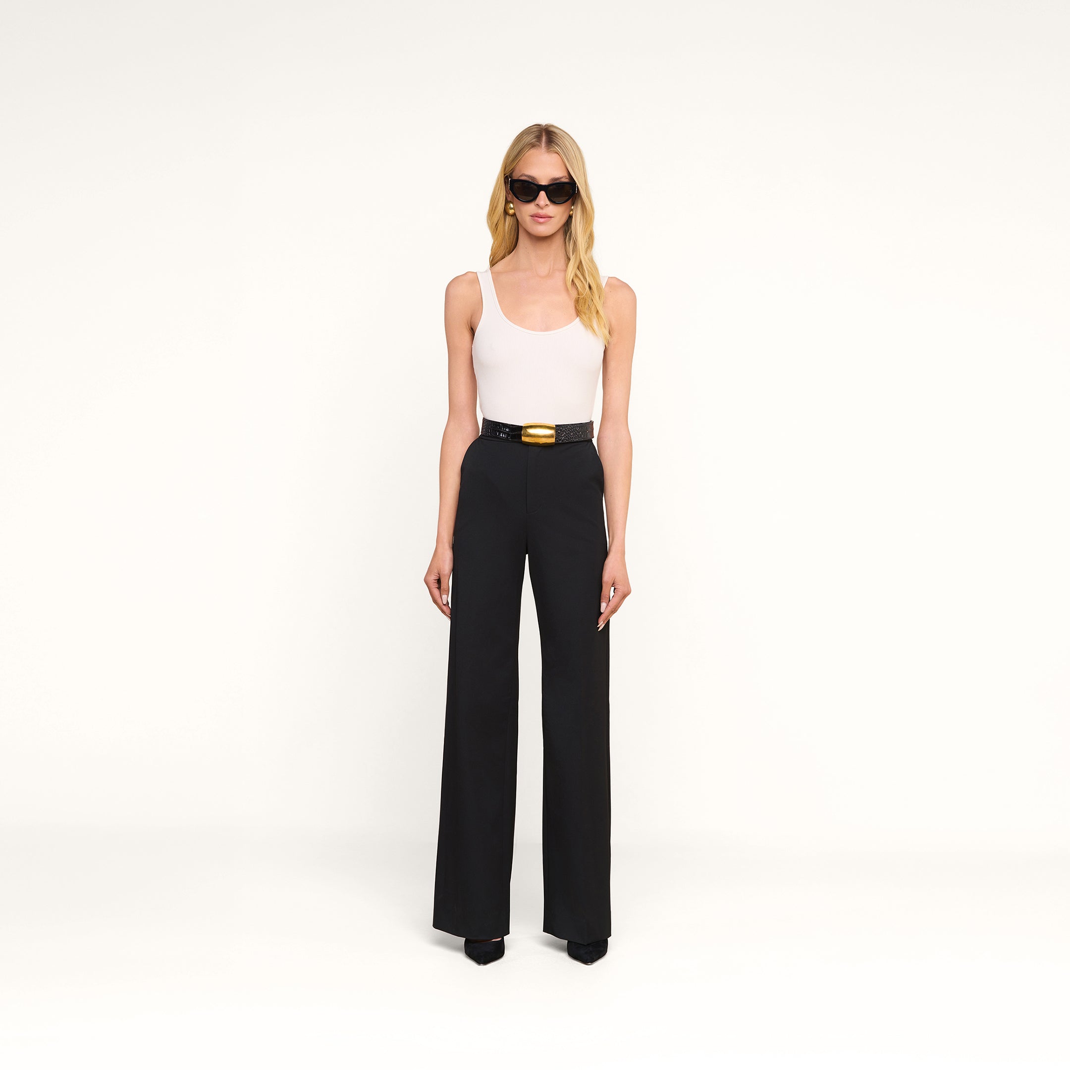 Une femme portant des lunettes de soleil, un débardeur blanc et le pantalon Livvy — un pantalon noir élégant à coupe droite, agrémenté dune ceinture noire dotée dune boucle dorée — se tient de face devant un fond beige. Une tenue décontractée, idéale pour passer du bureau au restaurant.