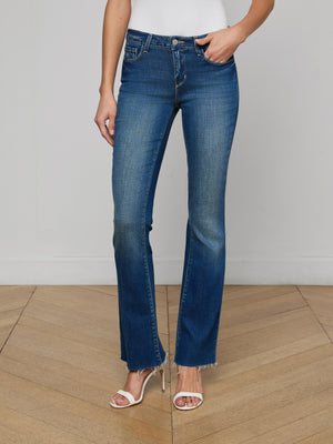 Someone stands on a wooden floor wearing Sneeki Low-Rise Straight-Leg Jeans in blue with a raw hem and white high-heeled sandals, one hand in their pocket, set against a plain light-colored background.