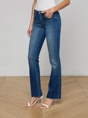 A person wears the Sneeki Low-Rise Straight-Leg Jean in blue stretch denim with a raw hem, paired with a white tank top and white high-heeled sandals, standing on a wooden floor, shown from shoulders down.