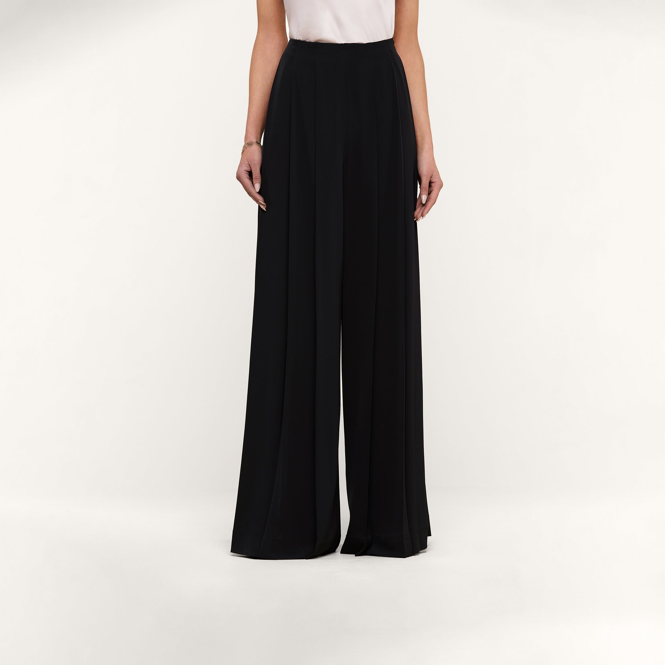 A person stands against a plain white background, showcasing the lower half of their body dressed in the Nova Pleated Wide-Leg Pant—a tailored, high-waisted black trouser—paired with a sleeveless white top.
