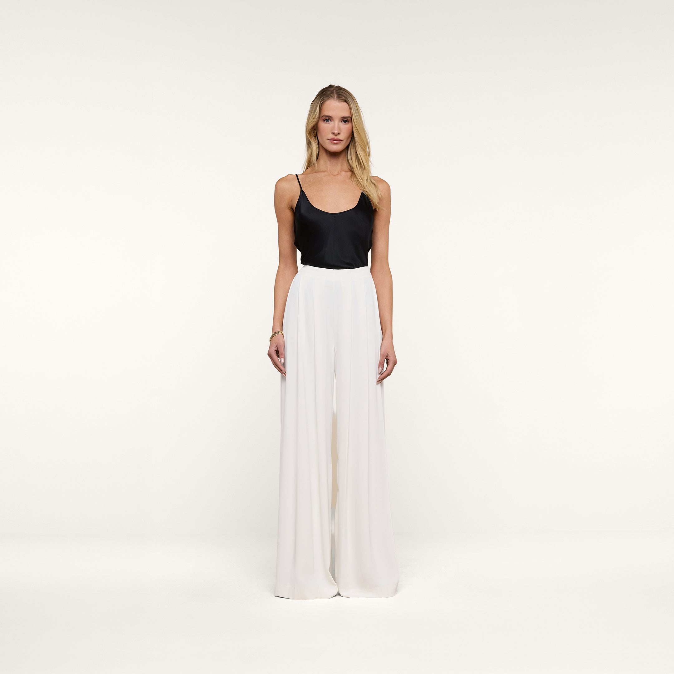 A woman stands facing forward against a plain white background, wearing a black sleeveless top paired with the Nova Pleated Wide-Leg Pant in ivory.