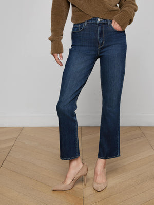 A person wears L’AGENCE’s Mira Cropped Bootcut Jean in dark blue stretch denim, paired with a brown sweater and beige heels, standing on a light wood herringbone floor against a white wall.