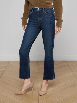 A person wearing a brown sweater, L'AGENCE Mira Cropped Bootcut Jean in dark blue stretch denim, and beige high-heeled shoes stands on a light wooden floor against a plain light-colored wall.