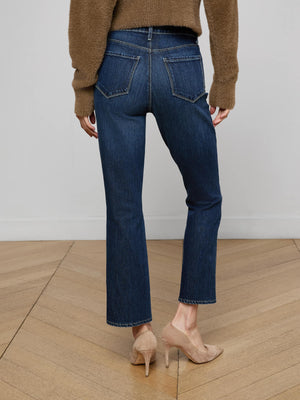 A person stands on a wood floor, facing away from the camera, wearing a brown sweater, L'AGENCE Mira Cropped Bootcut Jean in dark blue stretch denim, and beige high-heeled shoes.