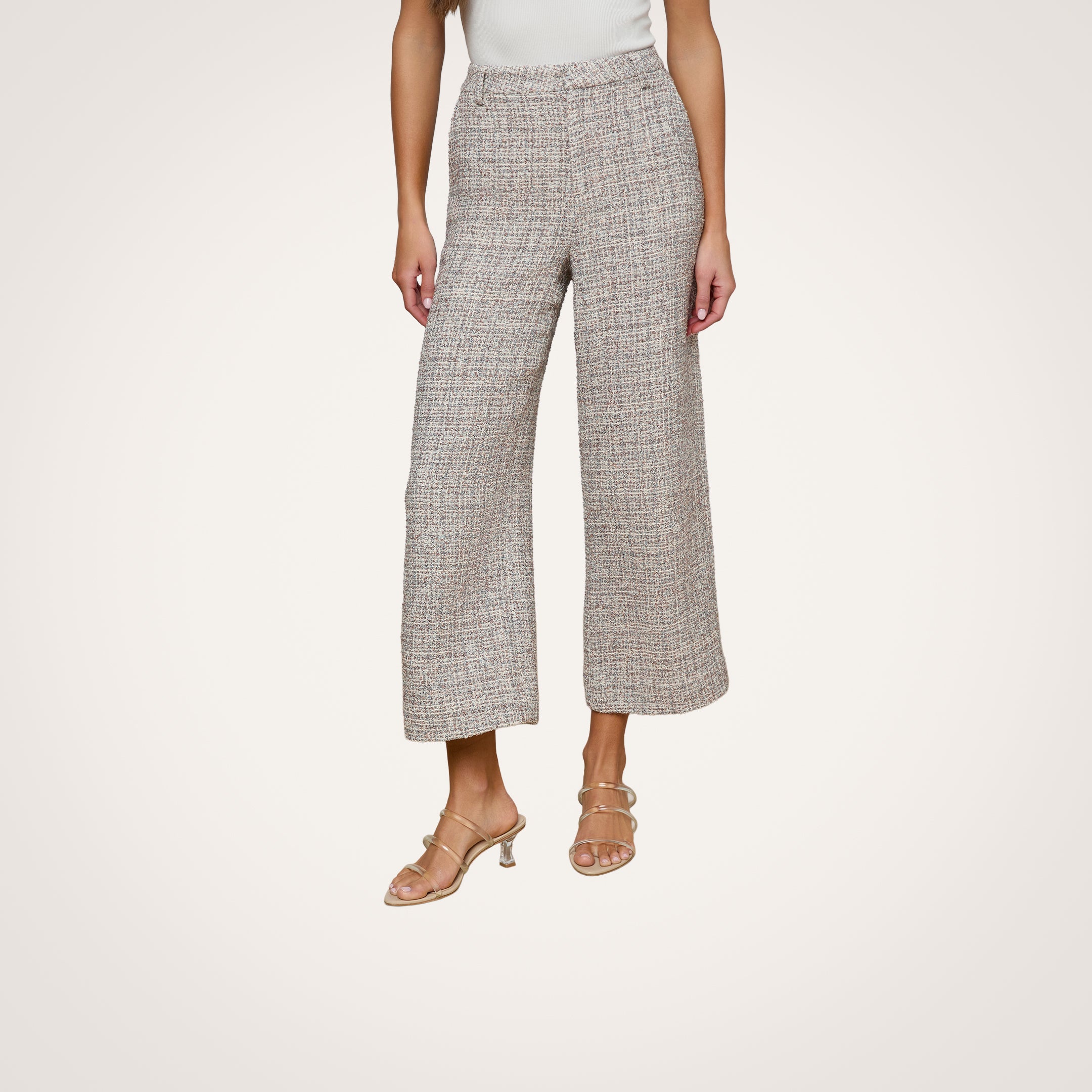 A woman models the Lula Cropped Tweed Trouser, a pair of wide-leg Italian tweed pants, along with beige heeled sandals. She stands against a plain white background, wearing a light-colored sleeveless top. Her head is not visible.