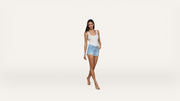 A woman with long dark hair, wearing a white tank top, light blue denim shorts, and beige shoes, stands against a plain white background with her hands in her pockets.