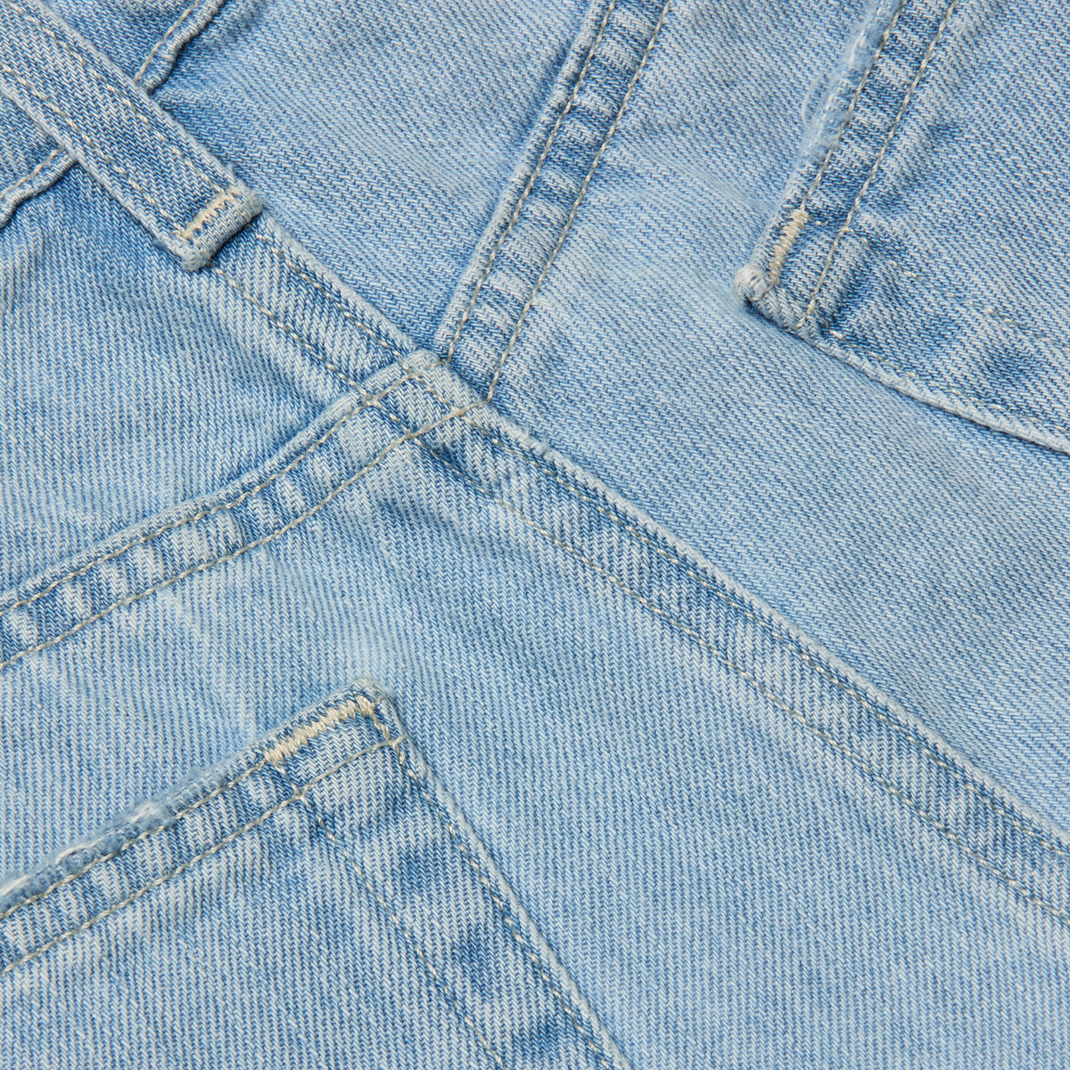 Close-up of the Teya Low-Rise Jean Short in a light blue wash, showcasing overlapping seams, pockets, and visible stitching details. The image highlights the texture, color, and structure of the denim material.