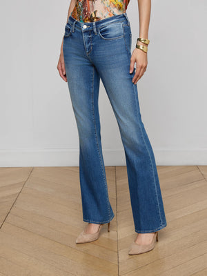 A person in L'AGENCE Cara Low-Rise Bootcut Jeans, a colorful top, gold bracelets, and beige high heels stands on a light wood floor against a plain white wall.