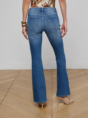 A person stands on a wooden floor wearing L'AGENCE Cara Low-Rise Bootcut Jeans, nude heels, a printed top, and a chunky gold bracelet. View is from the back, highlighting the jean's back pockets and subtle fading.