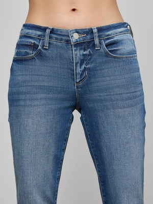 A close-up shows a person wearing the L'AGENCE Cara Low-Rise Bootcut Jean in blue stretch denim with button and zipper closure, highlighting the waistband and upper thighs against a plain background.