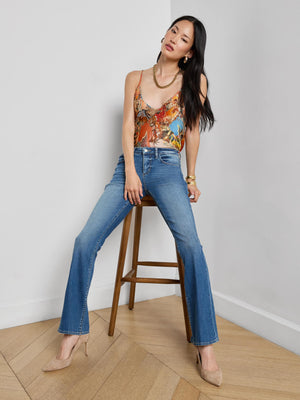 A woman with long dark hair poses confidently on a wooden stool against a white wall, wearing the L’AGENCE Cara Low-Rise Bootcut Jean, a colorful sleeveless top, beige pointed heels, and gold jewelry.