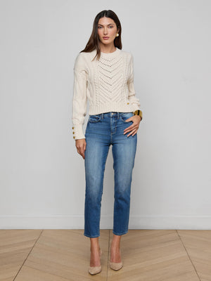 A woman with long brown hair wears the Celestia Slim-Leg Jean, a cream knit sweater, and beige heels, standing indoors against a light gray wall with neutral makeup and a confident posture.