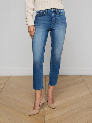 A person wearing the Celestia Slim-Leg Jean in stretch denim, a cream sweater with gold buttons, and beige heels stands on a wooden floor against a plain light gray wall.