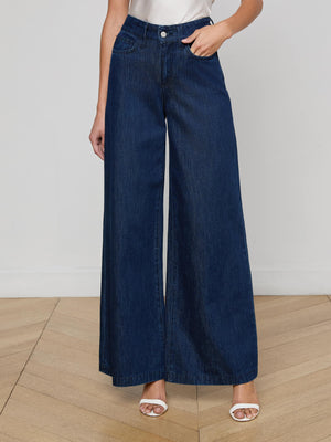 Wearing the Pacey Wide-Leg Jean in a dark wash, paired with white heeled sandals and a sleeveless white shirt, someone stands on a light wooden floor with one hand in their pocket.